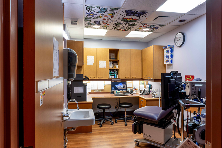 A small medical exam room with wooden cabinets, a cushioned examination chair, and two stools. A computer on the desk shows medical software. The ceiling features colorful art panels. A wall clock reads ten-thirty. The room appears clean and organized.